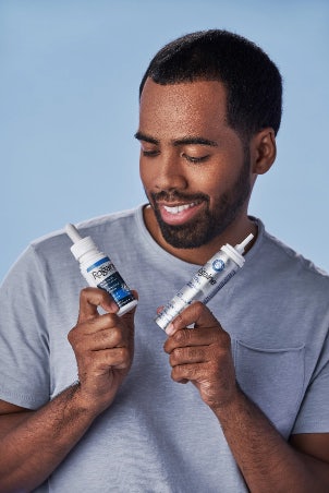 Man with dark brown hair and beard in gray t-shirt holding Rogaine Men's Minoxidil Foam and Solution