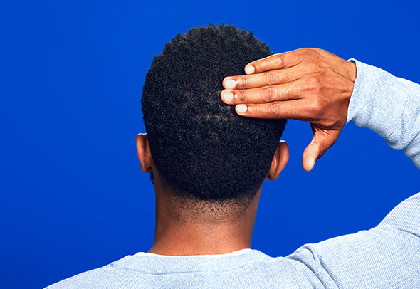 A man with dark brown hair touching the back of his head, on a blue background.  