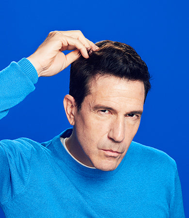 A man with brown hair and a blue sweater touching his hair.