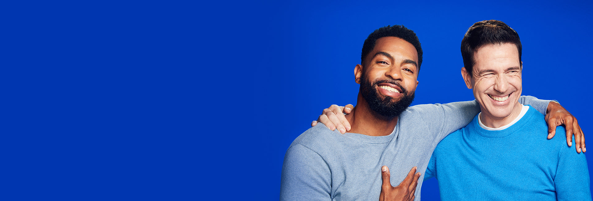 Two men in blue sweaters smiling on a blue background.